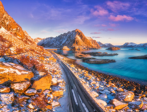 Aerial View Of Beautiful Mountain Road Near The Sea, Mountains, Purple Sky At Sunset In Lofoten Islands, Norway In Winter. Top View Of Road, Car, High Snowy Rocks With Stones, Coastline, Blue Water