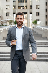 Photo closeup of caucasian entrepreneur or director man 40s in gray suit, walking down stairs of modern business center with takeaway coffee in hand