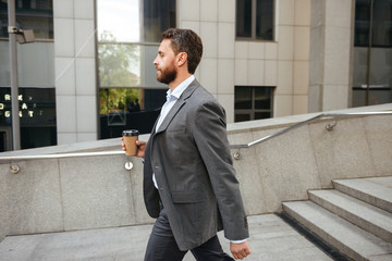 Profile photo of successful entrepreneur or director man 40s in gray suit holding takeaway coffee, and walking along street with modern business center background