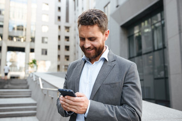 Photo of adult handsome man in gray suit typing or reading text message in mobile phone, while...