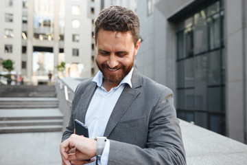 Photo of business man 40s in gray suit holding cell phone, and looking at smartwatch, while having break after working in office