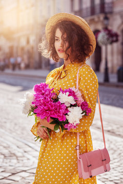 Outdoor portrait of young beautiful girl wearing yellow polka dot dress, straw hat, with small shoulder pink bag, holding peonies, walking in street of european city. Spring, summer fashion concept