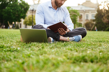 Photo of caucasian man wearing white shirt, writing down notes in notebook while sitting on grass...