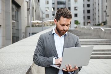 Image of business man 40s in formal wear holding open silver laptop, while standing in front of office building in urban area