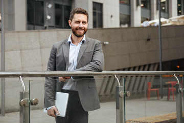 Image of successful entrepreneur man 40s in formal wear holding closed silver laptop in hand, while standing in front of business center