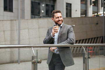 Photo of bearded business man 40s in gray suit looking aside with smile, while standing and drinking takeaway coffee in front of modern business center
