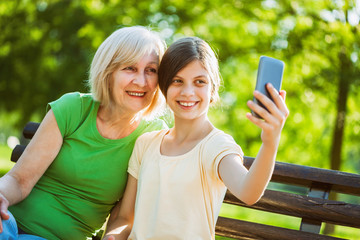 Grandmother and granddaughter are sitting in park and taking selfie. 