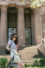 attractive girl in sunglasses sitting on bike and using smartphone near beautiful building with columns and stairs