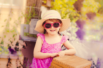A little girl wearing a sunhat and sunglasses is wearing a rattan suitcase and looking at the camera