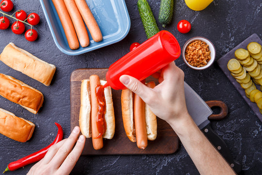 Picture on top of table with ingredients for hot dogs, cutting board, man's hands