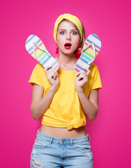 Young redhead girl in yellow t-shirt and blue jeans holding a summer flip flops sandals on pink background.