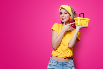 Young redhead girl in yellow t-shirt and blue jeans holding supermarket basket on pink background