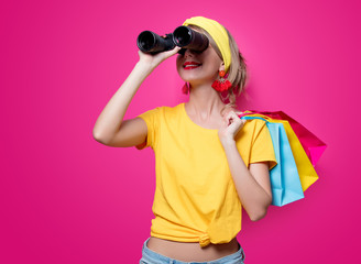 Young redhead girl in yellow t-shirt and blue jeans holding colorful bags and binoculars on pink...
