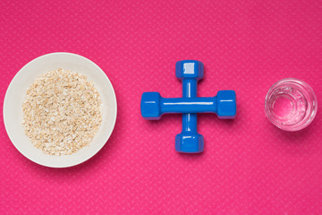 on a pink rug a plate with muesli, dumbbells lined with a plus sign and a glass with water, a concept, a sport and a healthy lifestyle, a top view