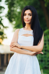 Vertical portrait of beautiful young female with dark hair, keeps hands crossed, has pleased expression, wears white dress, stands against blurred nature background, shows her natural beauty