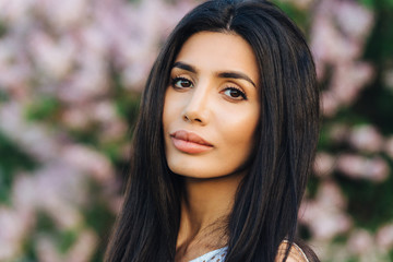 Close up portrait of pretty brunette young female with long dark hair, has serious look at camera, poses against blossom lilac blurred background outdoor, spends free time in botanical garden.