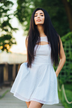 Confident pretty brunette female with serious look, make up and clean skin, wears festive white dress, strolls in park at city, keeps hands in pockets, likes to be photographed. People and leisure