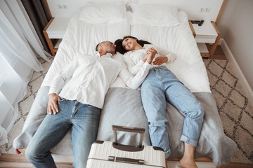 Photo from above of joyful couple man and woman in casual jeans clothing, resting together and lying in bed at hotel room with big luggage. Holiday or vacation concept