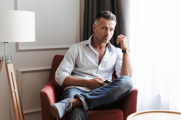 Photo of caucasian handsome man 30s in casual wear using smartphone, while sitting in armchair in living room or hotel apartment