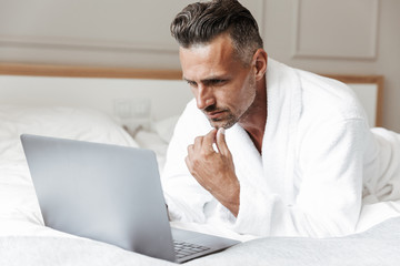Photo of european man with gray beard wearing white bathrobe using silver laptop, while lying on bed in bedroom or hotel apartment