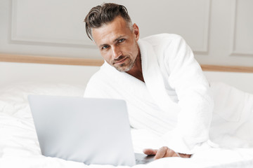 Image of european unshaved man with gray beard wearing white bathrobe looking at camera, while...