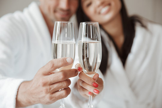 Blurry image closeup of caucasian handsome man and beautiful asian woman, holding and clinking glasses with champagne at hotel room
