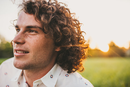 Handsome young freckled male smiling with white toothy smile and curly hair posing on the lawn in the park looking a side, sunset view. Copy space for your advertising. People, lifestyle and emotion