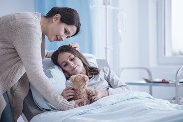 Caring mother giving a plush toy to sick daughter lying in a hospital bed - obrazy, fototapety, plakaty