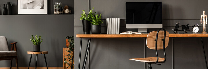 Wooden desk with fresh plants, mockup computer, books and decor standing in dark open space living...