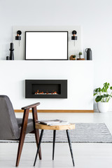 Grey armchair next to table in living room interior with mockup of poster above fireplace. Real photo