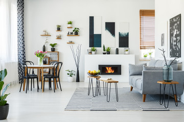Black chairs at dining table in bright living room interior with grey sofa near fireplace. Real...