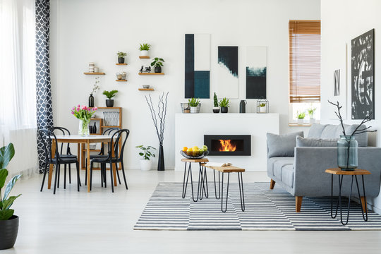Black chairs at dining table in bright living room interior with grey sofa near fireplace. Real photo