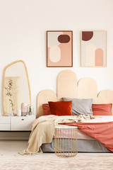 Real photo of a king-size bed standing next to a shelf with a mirror in a bedroom interior with two posters on a wall