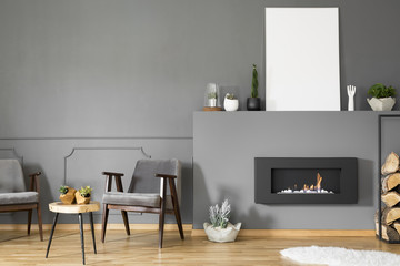 Fireplace under empty poster with mockup in grey living room interior with armchairs. Real photo