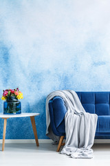 Comfortable dark blue settee with gray blanket and small side table with fresh flowers in a vase against blue and white ombre wall in a living room interior. Copy space background. Real photo. © Photographee.eu