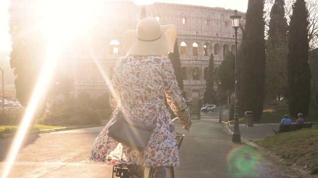 Beautiful young woman in colorful fashion dress with bike walking toward colosseum in Rome at sunset with trees happy attractive tourist girl in colle oppio
