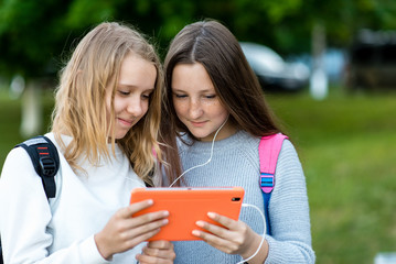 Beautiful little two girls. Summer in nature. In her hands holds a tablet. Watching video listening music. Looks social network. Emotional happy rejoices. The best girlfriends in nature after school.