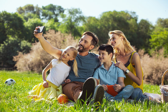 Say cheese. Inspired well-built man smiling and taking selfies with his family in the open air