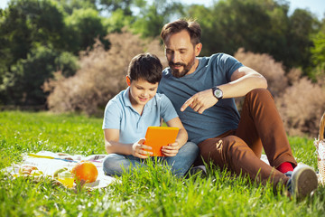 Our leisure. Content lovely boy holding a tablet while sitting with his father in the park