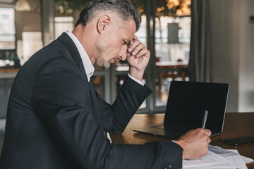 Image of handsome caucasian businessman wearing white shirt and black suit sitting at table in office, during work with documents and laptop