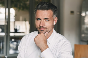 Obraz premium Portrait closeup of successful adult man 30s wearing white shirt looking at camera and touching chin, while working in office using white earbud