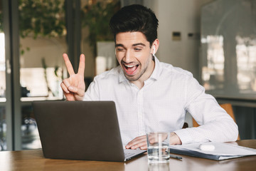 Portrait of joyful office man 30s wearing white shirt laughing and showing peace sign at laptop, during video conference or call using bluetooth earbud