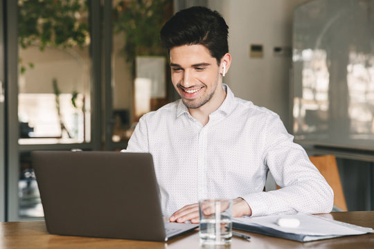 Image of handsome businessman wearing white shirt and modern earpod smiling while sitting at table in office, and looking at laptop