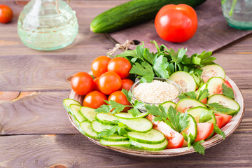 Fresh slices of cucumbers, tomatoes, sesame seeds in a bowl and parsley leaves on a plate on a wooden table