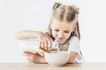 happy little schoolgirl pouring milk into cereal for breakfast isolated on white