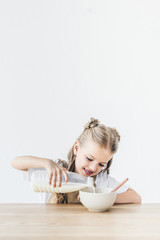 smiling little schoolgirl pouring milk into cereal for breakfast isolated on white