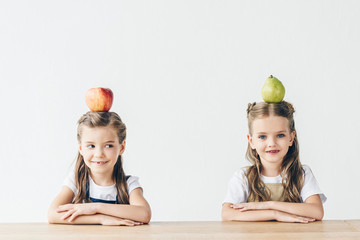 adorable little schoolgirls with apple and pear on heads sitting at table isolated on white
