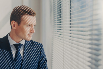 Fototapeta premium Indoor shot of pensive male economist dressed in formal outfit, looks pensively at window with jalousie conemplates about work during dinner break feels tired after working. Business, people, thoughts