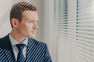 Fototapeta premium People and career concept. Young entrepreneur in rich suit, thinks about future of his company, stands near office window, has concentrated thoughtful look. Male managing director indoor in cabinet