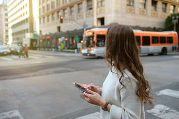 Beautiful young woman on the boulevard in urban scenery, downtown, at sunset, holding smartphone